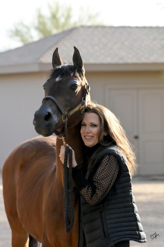 woman with long brown hair, wearing a black vest with a black, and tan long sleeved top is holding the halter of a bay Arabian horse with a small star, and putting their heads close together and looking at the camera.